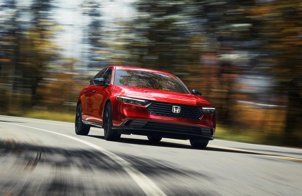 2024 red Honda Accord Hybrid driving on a winding road lined with blurred autumn trees.