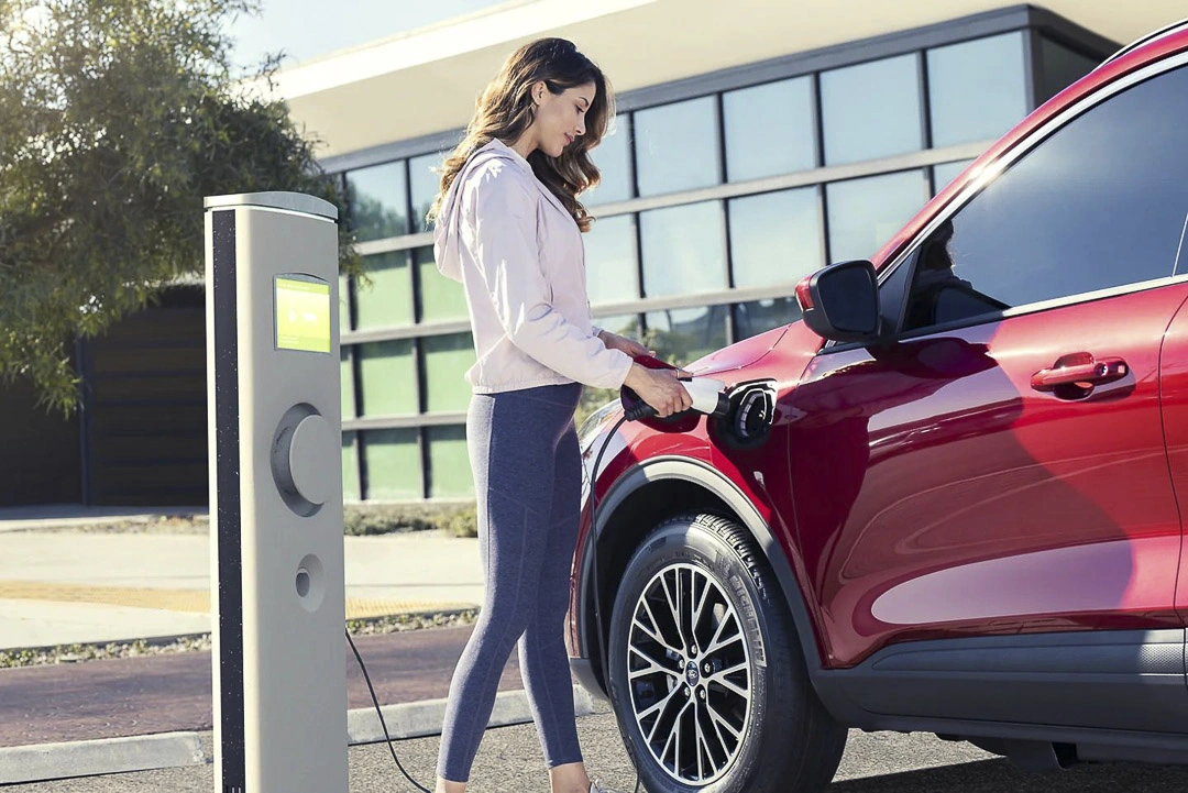 Woman plugging in a 2022 red Ford Escape PHEV at a modern residential charging station during the day.