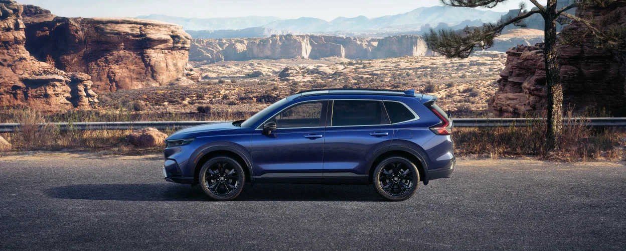 2023 blue Honda CR-V Hybrid side view parked in a desert landscape with rock formations and clear sky.