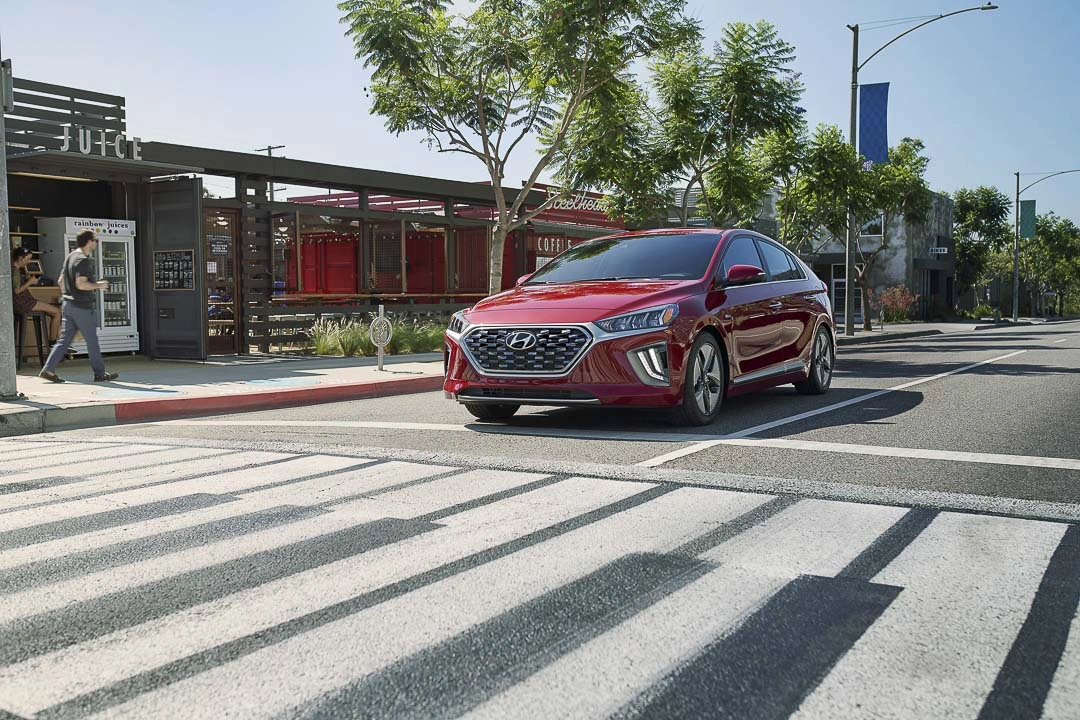2022 red Hyundai Ioniq Hybrid crossing a pedestrian crosswalk in a modern urban area with cafes and shops in the background.