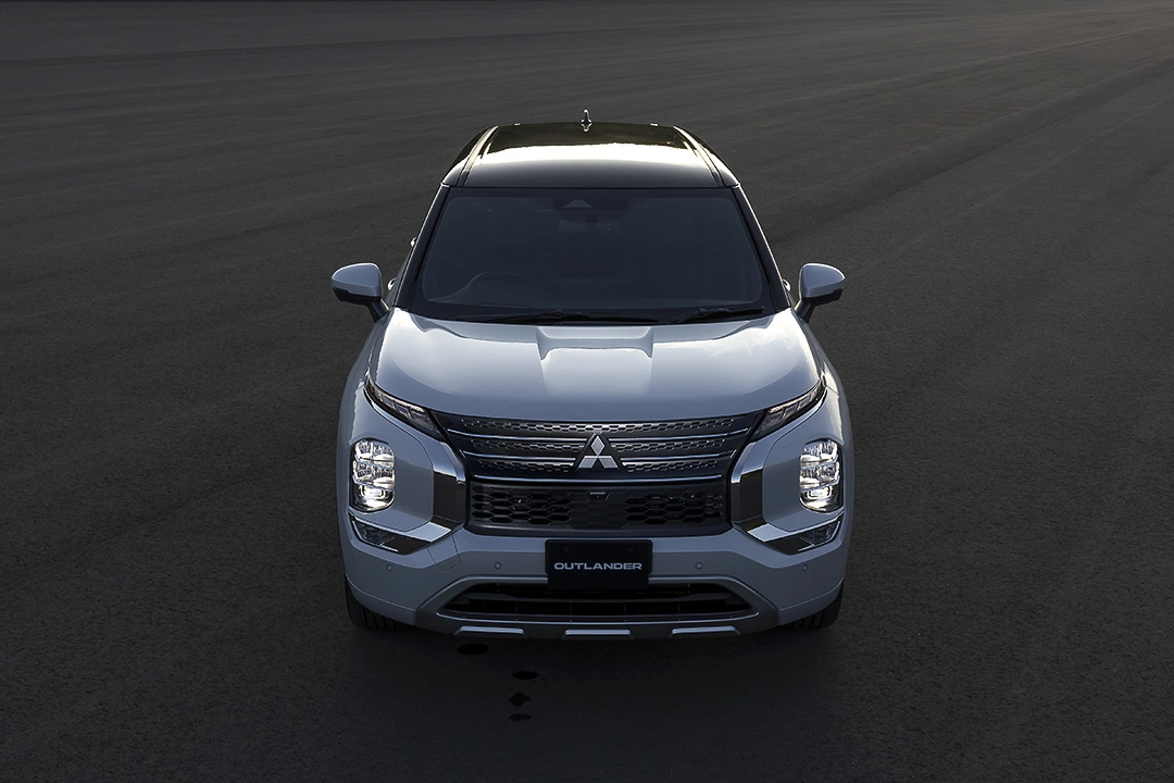 Front view of a 2022 silver Mitsubishi Outlander PHEV parked on a large dark asphalt surface, headlights on and Mitsubishi logo centered on the black grille.
