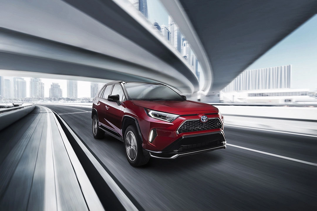 2021 red Toyota RAV4 Prime driving on an urban highway under a modern viaduct, dynamic silhouette and skyscrapers in the background.