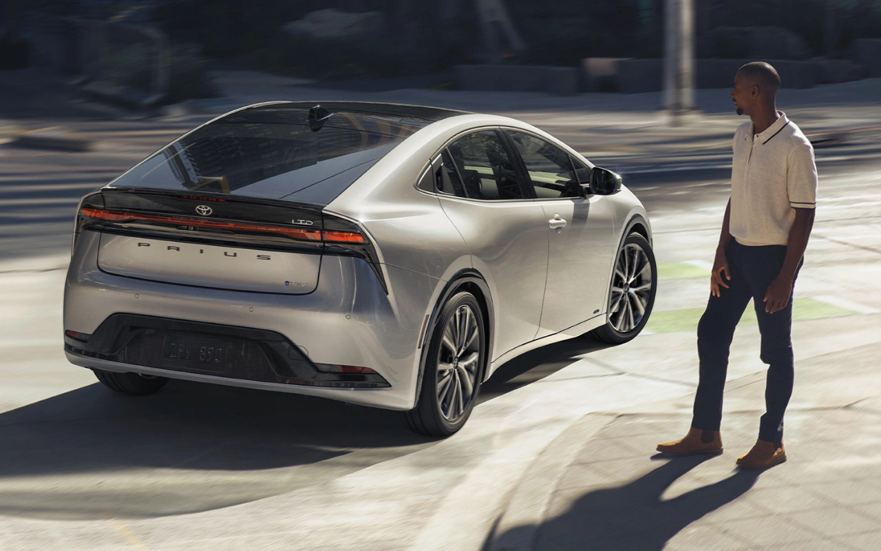2024 silver Toyota Prius Hybrid rear view driving in the city, man watching the car at a crosswalk, distinctive futuristic design.